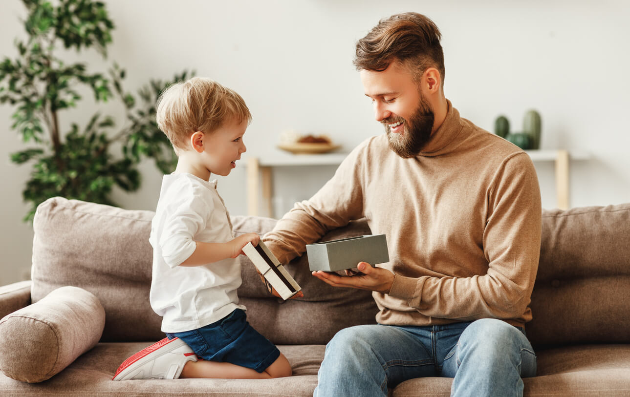Excited young man sitting on sofa and opening gift box given by cute little boy during home celebration of Fathers day father's day gifts for outdoorsy dad