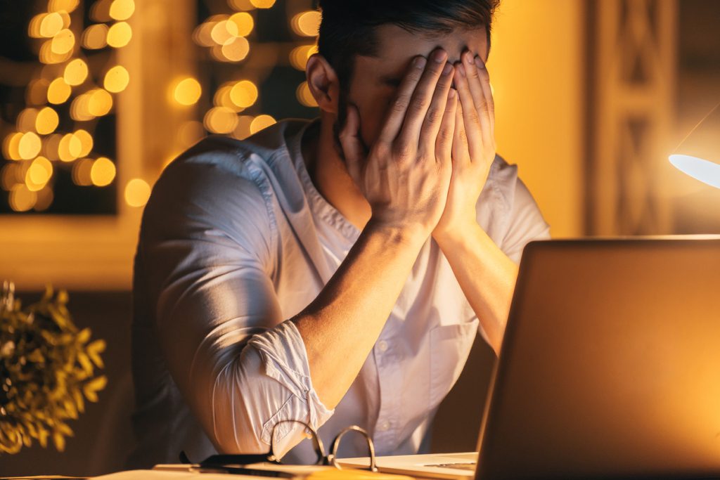 Frustrated young man covering face with hands while sitting at his working place at night time with Christmas lights in the background Work Life Balance During the Holiday Season