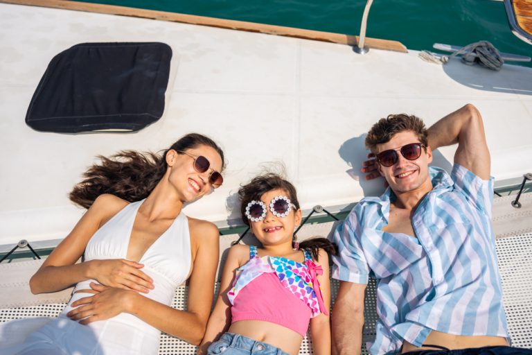Book summer travel early 2026 wave season deals Caucasian attractive family lying down on deck of yacht while yachting. Young beautiful couple hanging out and spend time with daughter while catamaran boat sailing during holiday summer trip together