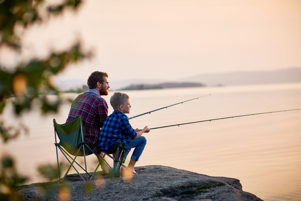 Side view portrait of father and son sitting together on rocks fishing with rods in calm lake waters with landscape of setting sun, both wearing checkered shirts, shot from behind tree fishing with kids