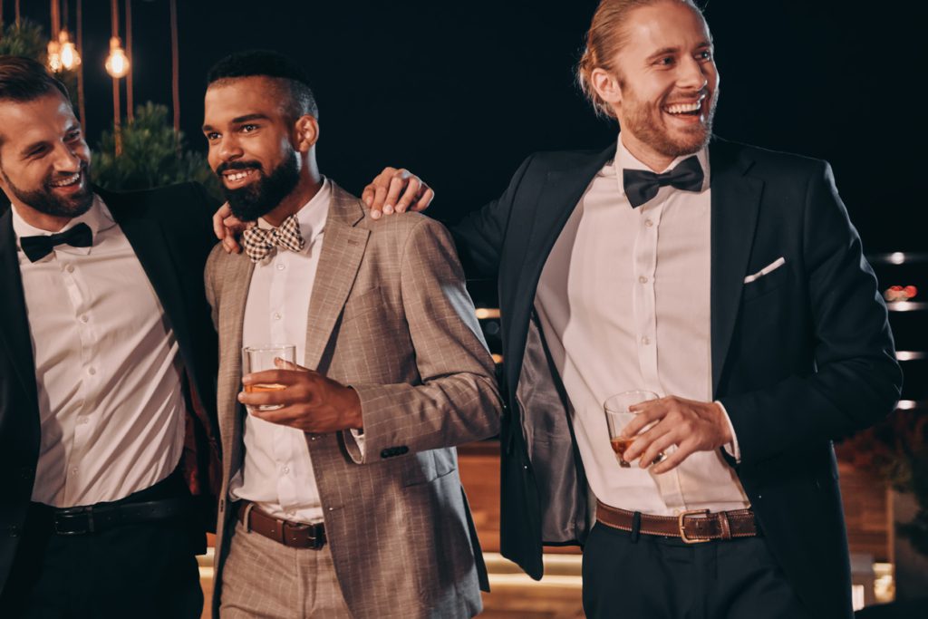 Three handsome men in suits bonding and drinking whiskey while spending time on party male wedding guest what to wear to a wedding