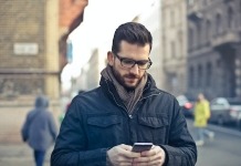 Man Wearing Black Zip Jacket Holding Smartphone Surrounded by Grey Concrete Buildings - best men's spring jackets