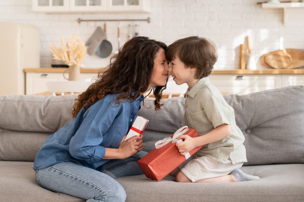 Loving mom and son sit bonding on couch at home hold gifts in hands. Cute mother and adorable small kid celebrate happy family day exchanging presents for holiday event cheerful and excited together sentimental gifts