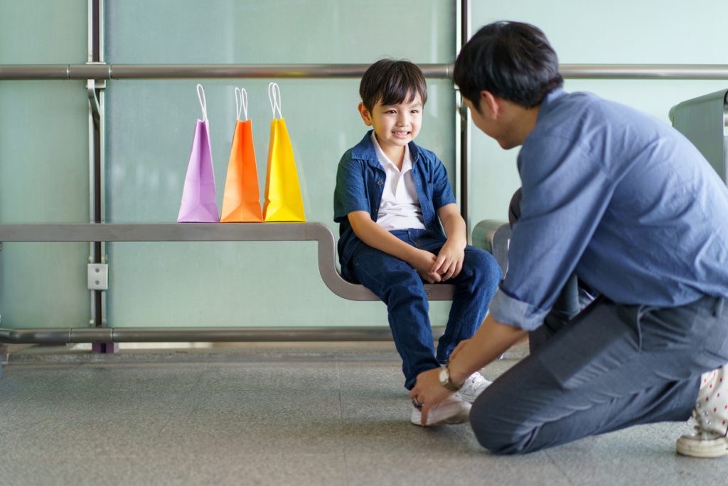 back-to-school shopping day - An Asian man is tying his son's shoelaces as the boy sits on a bench while they wait for the train at the railway station.