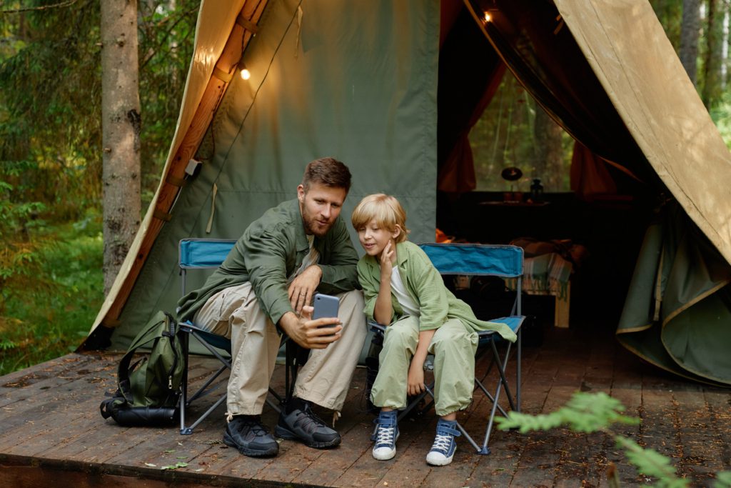 Father and son sitting on chairs outside their tent in forest, taking a selfie together. Smiling and bonding in natural surroundings, tent lit with cozy lights