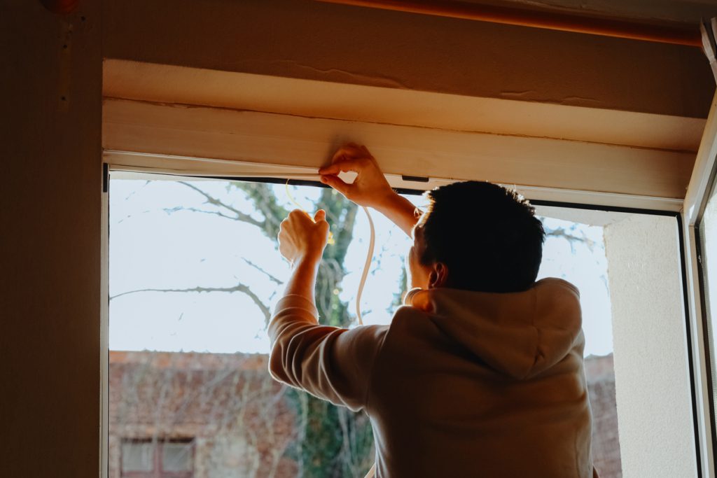 Side close-up portrait of one young Caucasian man standing from the back, gluing special rubber to the top of an interior window frame to seal cracks for the winter in a second floor room. The concept of repair and insulation of window frames. Home Maintenance Checklist for Winter 